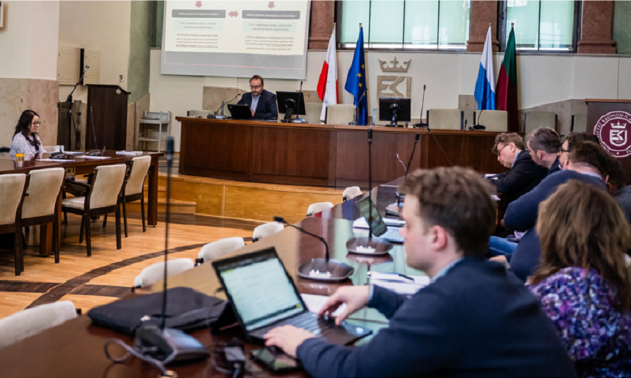 Sala na której siedzą ludzie w garniturach z komputerami. Z tyłu rzutnik i prezentacja oraz mężczyzna siedzący na środku sali obok niego flaga Polski i Unii Europejskiej.