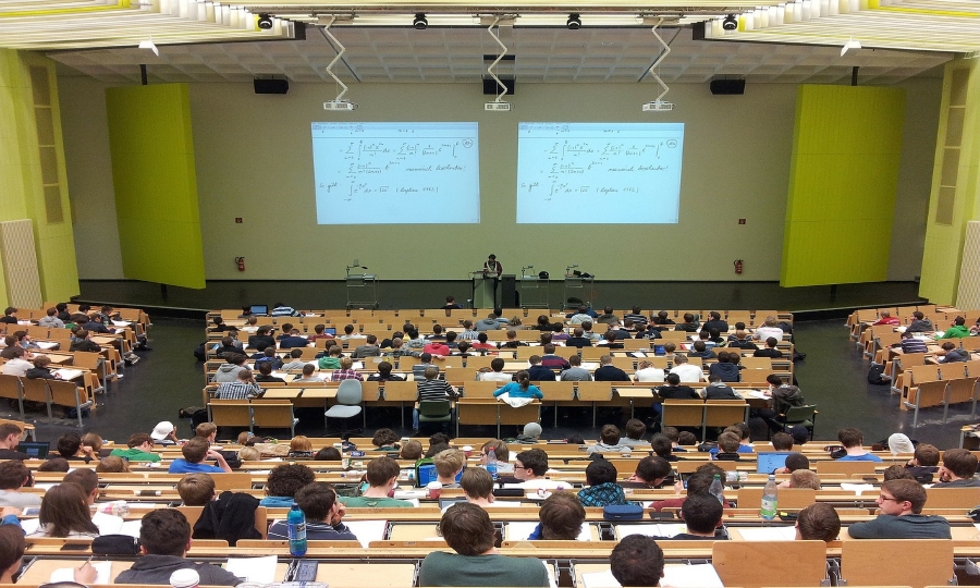 Zdjęcie przedstawia aulę wykładową na wyższej uczelni, na której siedzą studenci i słuchają wykładu prowadzącego.